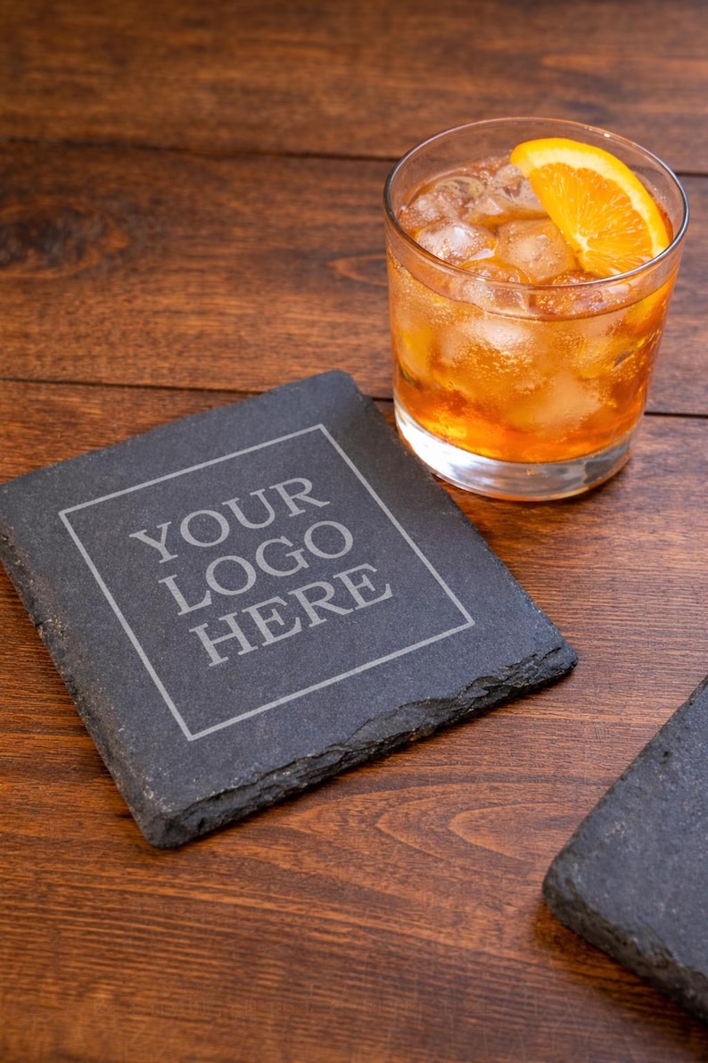 May include: A square, dark gray slate coaster with the text "YOUR LOGO HERE" in a white, outlined font. A glass of amber-colored drink with ice and an orange slice sits nearby. The coaster is on a dark wood surface.