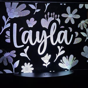 May include: A black illuminated sign with the name "Layla" in a cursive font. The sign is decorated with holographic floral and leaf patterns. The light shines through the design, creating a glowing effect.