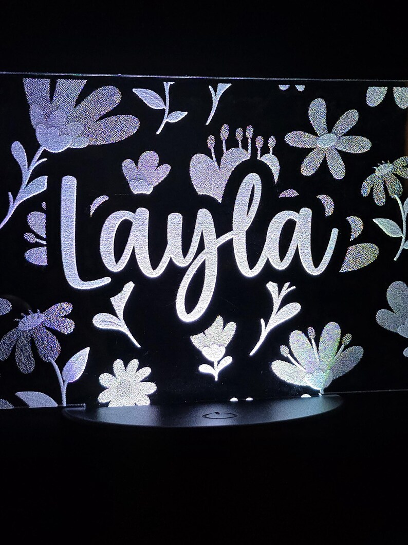 May include: A black illuminated sign with the name "Layla" in a cursive font. The sign is decorated with holographic floral and leaf patterns. The light shines through the design, creating a glowing effect.