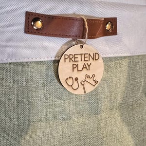 May include: A light green and blue fabric storage bin with a brown leather handle. A wooden tag with the words "PRETEND PLAY" and illustrations of a stethoscope, crown, and wand hangs from the handle.