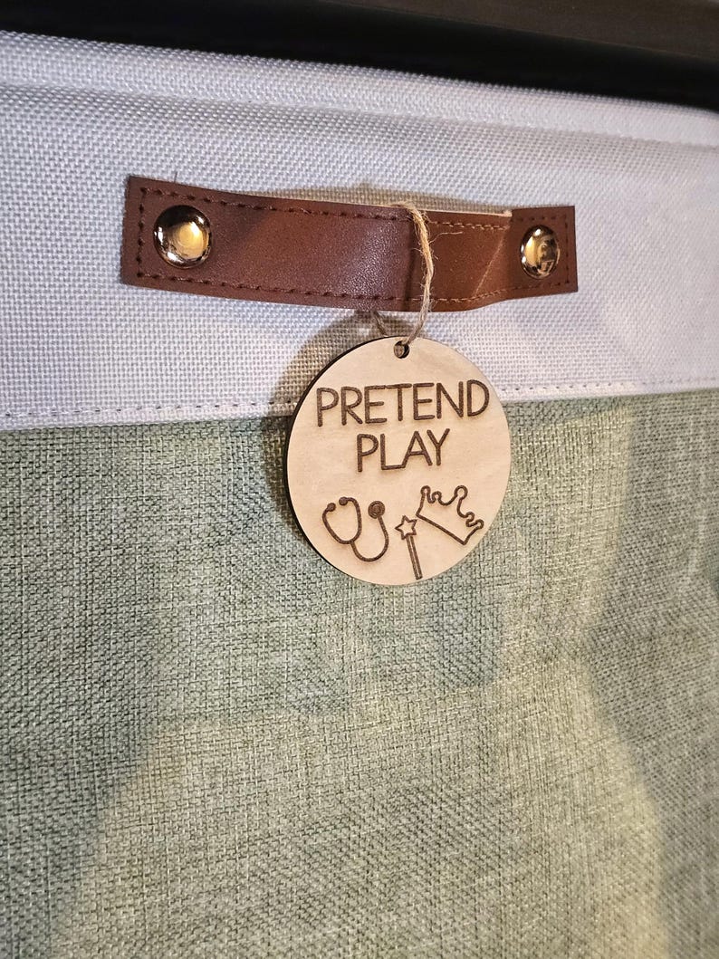 May include: A light green and blue fabric storage bin with a brown leather handle. A wooden tag with the words "PRETEND PLAY" and illustrations of a stethoscope, crown, and wand hangs from the handle.