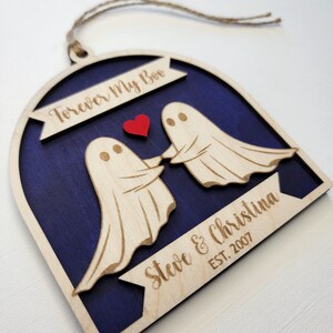 May include: A wooden ornament shaped like an arch with two ghosts holding hands and a red heart in the middle. The ornament is painted a dark blue color and has the text "Forever My Boo" at the top and "Steve & Christina EST. 2007" at the bottom.
