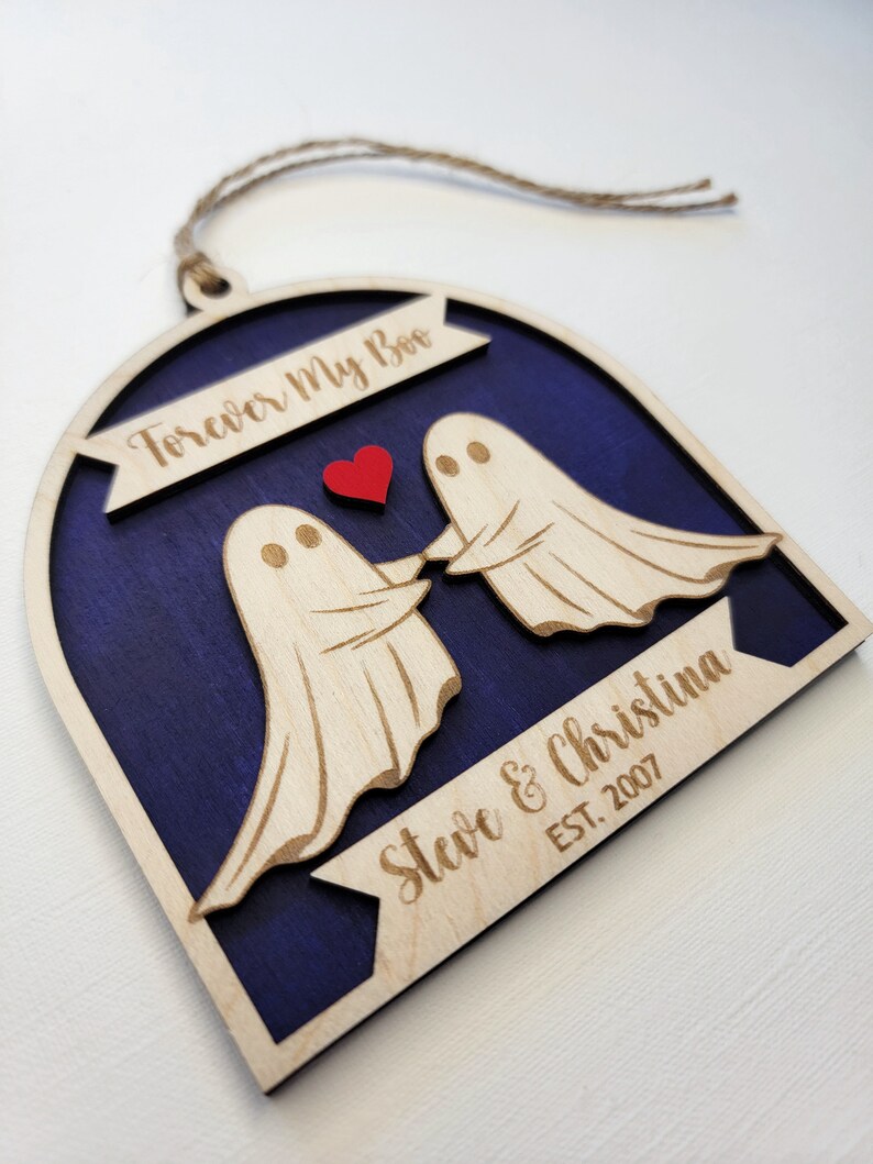 May include: A wooden ornament shaped like an arch with two ghosts holding hands and a red heart in the middle. The ornament is painted a dark blue color and has the text "Forever My Boo" at the top and "Steve & Christina EST. 2007" at the bottom.