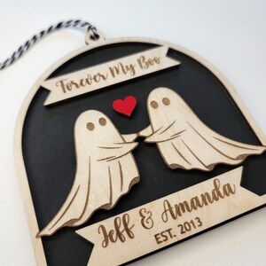 May include: A wooden ornament shaped like a ghost couple holding hands with a red heart between them. The ornament is black with a white border and has the text "Forever My Boo" at the top and "Jeff & Amanda EST. 2013" at the bottom.