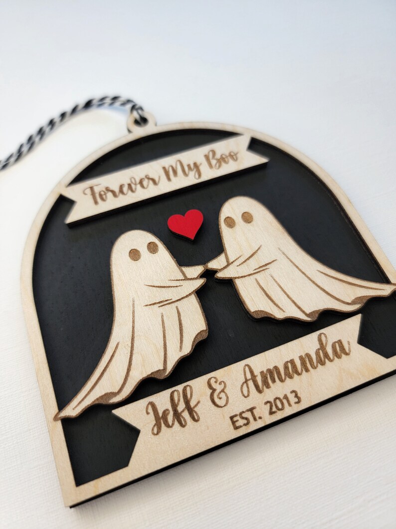 May include: A wooden ornament shaped like a ghost couple holding hands with a red heart between them. The ornament is black with a white border and has the text "Forever My Boo" at the top and "Jeff & Amanda EST. 2013" at the bottom.