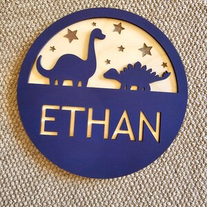 May include: A navy blue wooden circle with a cutout of two dinosaurs and stars. The name "ETHAN" is cut out in gold lettering.