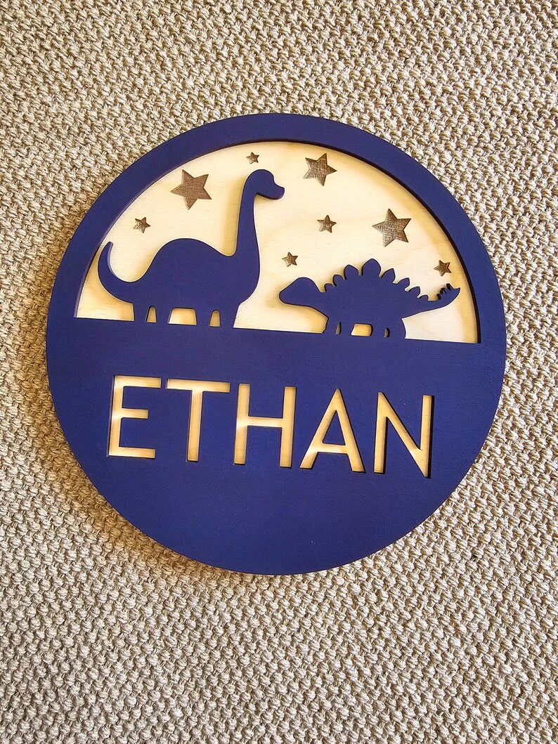 May include: A navy blue wooden circle with a cutout of two dinosaurs and stars. The name "ETHAN" is cut out in gold lettering.