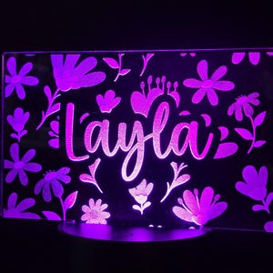May include: A purple LED night light with the name "Layla" in a cursive font, surrounded by floral designs. The light is rectangular and sits on a round base, creating a soft, ambient glow in a dark setting.