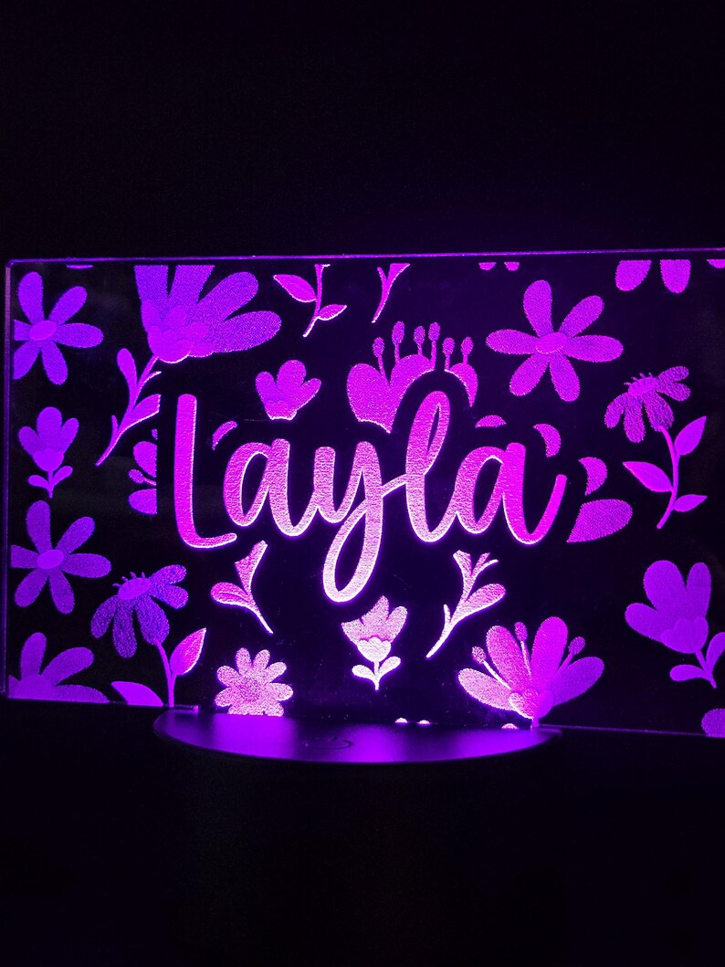 May include: A purple LED night light with the name "Layla" in a cursive font, surrounded by floral designs. The light is rectangular and sits on a round base, creating a soft, ambient glow in a dark setting.