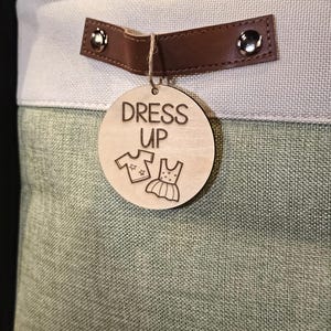 May include: A storage bin with a light green and gray fabric exterior. A brown leather handle is attached with silver rivets. A wooden tag with the words "DRESS UP" and clothing illustrations hangs from the handle.