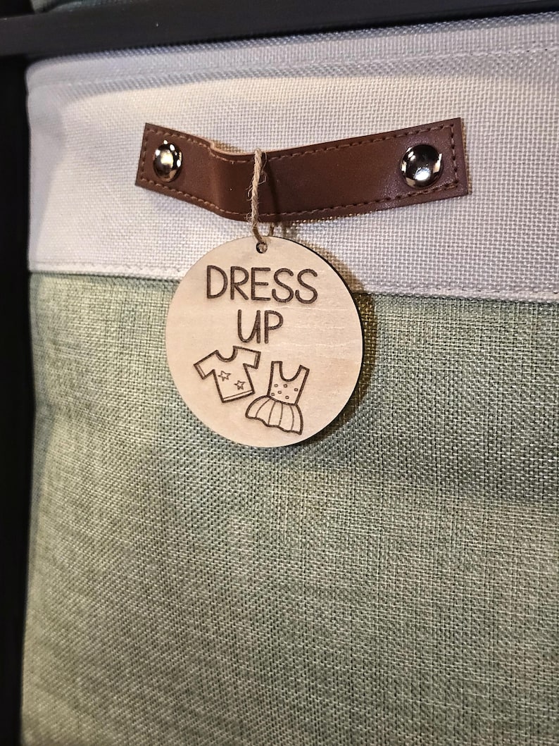 May include: A storage bin with a light green and gray fabric exterior. A brown leather handle is attached with silver rivets. A wooden tag with the words "DRESS UP" and clothing illustrations hangs from the handle.