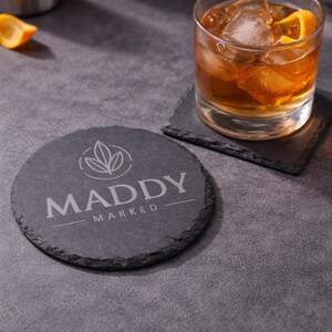 May include: A round, dark gray slate coaster with the text "MADDY MARKED" and a leaf design. A square coaster and a glass of amber-colored drink with ice and an orange peel are also visible.