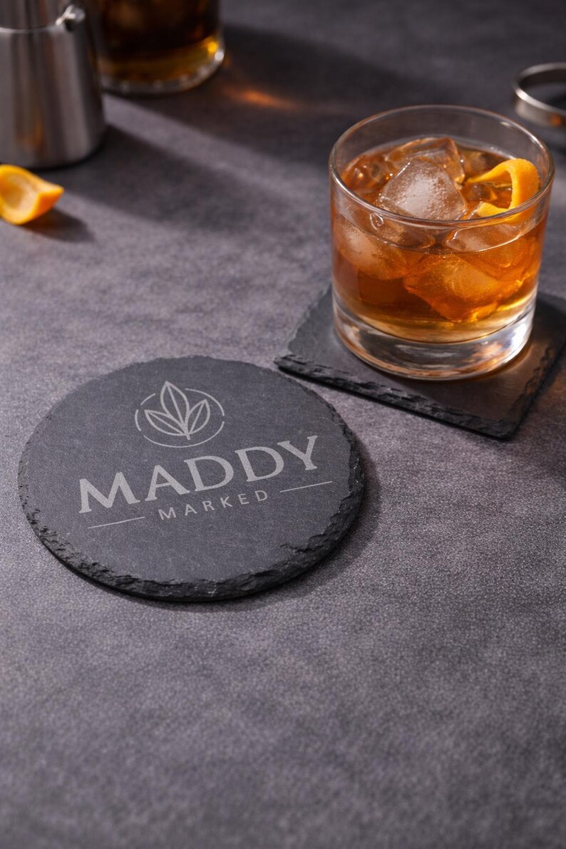 May include: A round, dark gray slate coaster with the text "MADDY MARKED" and a leaf design. A square coaster and a glass of amber-colored drink with ice and an orange peel are also visible.