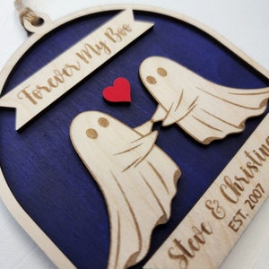 May include: A wooden ornament shaped like an arch with two ghosts holding hands. The ghosts are white and the arch is painted a dark blue. The text "Forever My Boo" is on the top of the arch and "Steve & Christine EST. 2007" is on the bottom. A red heart is between the ghosts.