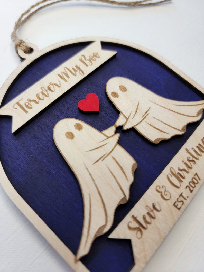 May include: A wooden ornament shaped like an arch with two ghosts holding hands. The ghosts are white and the arch is painted a dark blue. The text "Forever My Boo" is on the top of the arch and "Steve & Christine EST. 2007" is on the bottom. A red heart is between the ghosts.