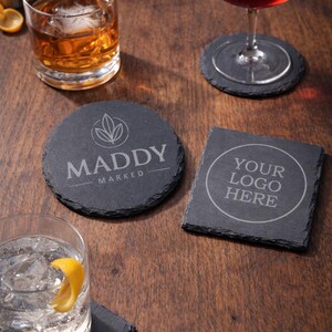 May include: A collection of black slate coasters with drinks on a wooden surface. One round coaster features the text "MADDY MARKED" and a leaf design. Another square coaster has the text "YOUR LOGO HERE".