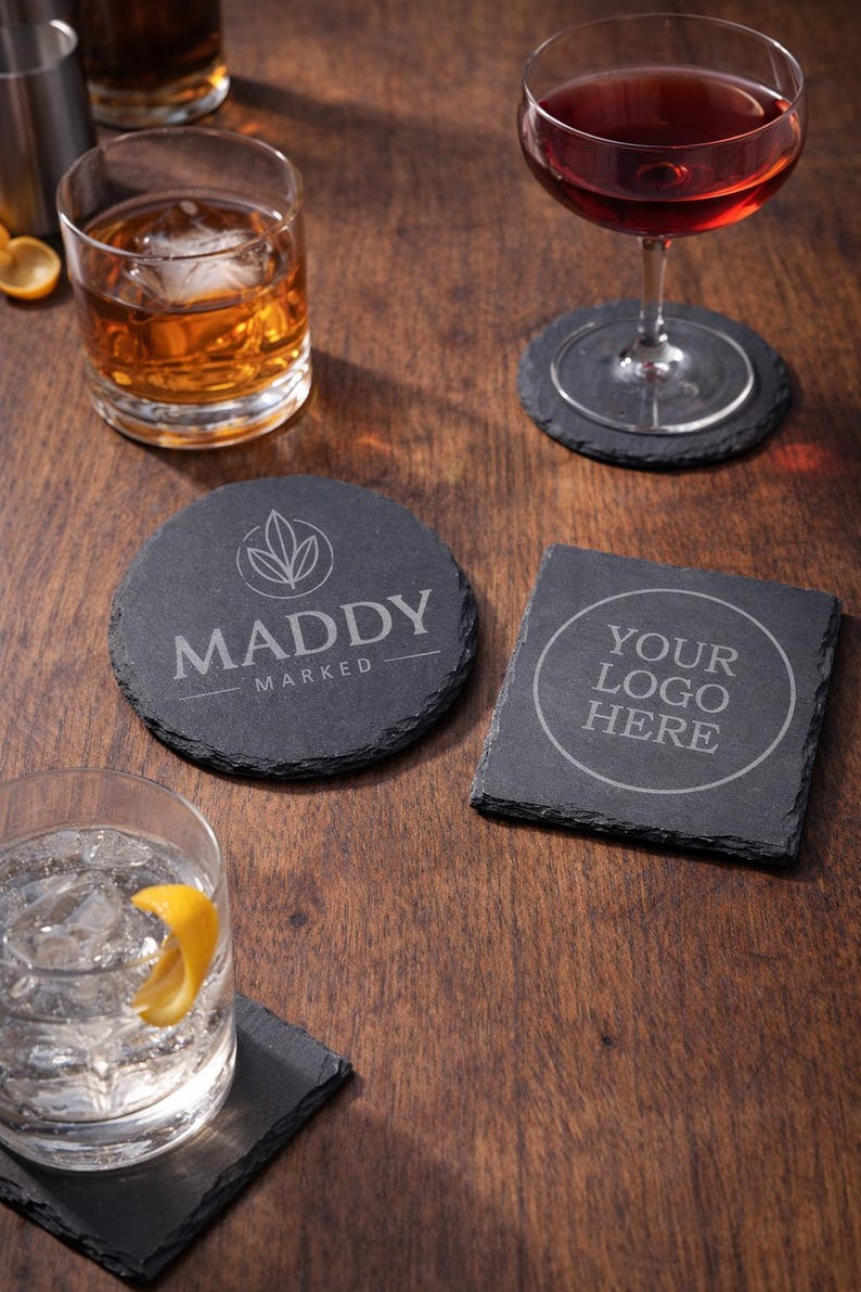 May include: A collection of black slate coasters with drinks on a wooden surface. One round coaster features the text "MADDY MARKED" and a leaf design. Another square coaster has the text "YOUR LOGO HERE".