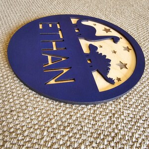 May include: A blue circular wooden sign with the name "ETHAN" cut out in the center. The sign features a silhouette of a dinosaur and stars on a light wood background.