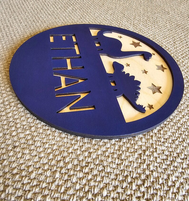 May include: A blue circular wooden sign with the name "ETHAN" cut out in the center. The sign features a silhouette of a dinosaur and stars on a light wood background.