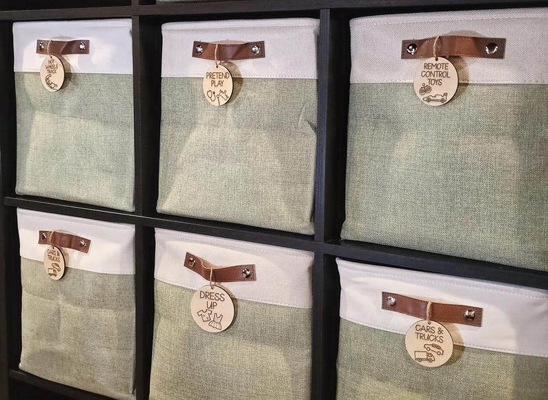 May include: Storage bins in a dark wood shelf. The bins are two-toned, with a light green lower section and a white upper section. Each bin has a brown leather handle and a wooden tag with text and illustrations. The tags read: "Hot Wheels Garage", "Pretend Play", "Remote Control Toys", "Cars & Trucks", and "Dress Up".