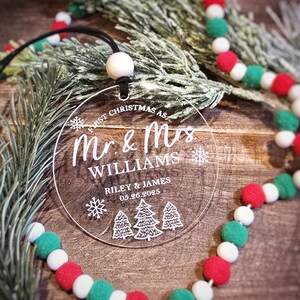 May include: Clear acrylic Christmas ornament with the text "First Christmas as Mr. & Mrs. Williams" and the date 05.26.2025. The ornament has a black string for hanging and is decorated with snowflakes and Christmas trees. A festive garland with red, white, and green pom-poms is in the background.