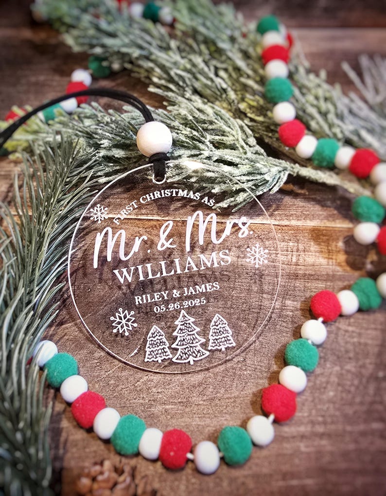 May include: Clear acrylic Christmas ornament with the text "First Christmas as Mr. & Mrs. Williams" and the date 05.26.2025. The ornament has a black string for hanging and is decorated with snowflakes and Christmas trees. A festive garland with red, white, and green pom-poms is in the background.