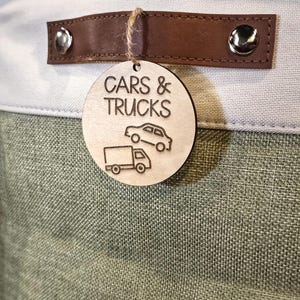 May include: A storage bin with a light green and white fabric exterior. A brown leather handle is attached with silver rivets. A wooden tag with the words "CARS & TRUCKS" and car and truck illustrations hangs from the handle.