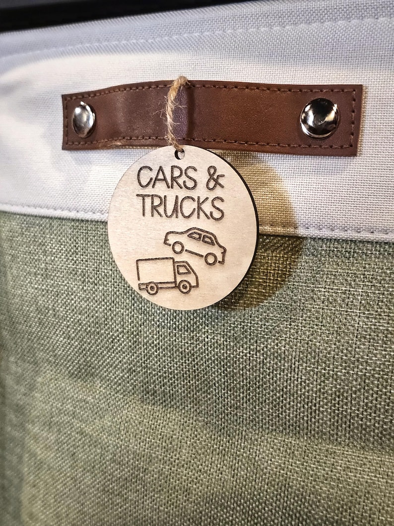 May include: A storage bin with a light green and white fabric exterior. A brown leather handle is attached with silver rivets. A wooden tag with the words "CARS & TRUCKS" and car and truck illustrations hangs from the handle.