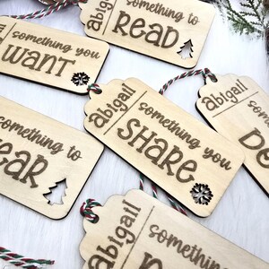 Pu&ograve; includere: Cinque etichette regalo in legno con il testo "abigail" e "something you" seguito da una parola diversa su ogni etichetta: "want", "read", "share", "need" e "wear". Ogni etichetta ha un piccolo ritaglio di albero di Natale.
