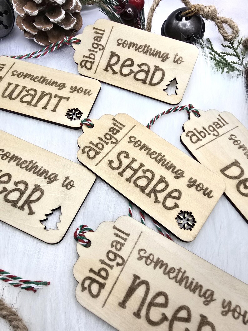 Pu&ograve; includere: Cinque etichette regalo in legno con il testo "abigail" e "something you" seguito da una parola diversa su ogni etichetta: "want", "read", "share", "need" e "wear". Ogni etichetta ha un piccolo ritaglio di albero di Natale.
