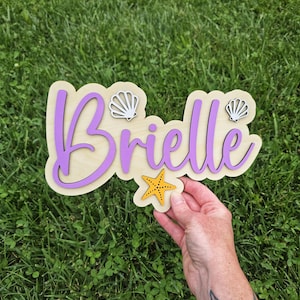 May include: Wooden sign with the name "Brielle" in purple script, outlined in a darker purple, on a light wood base. The sign is decorated with two white seashell cutouts and a yellow starfish. The sign is held over green grass.