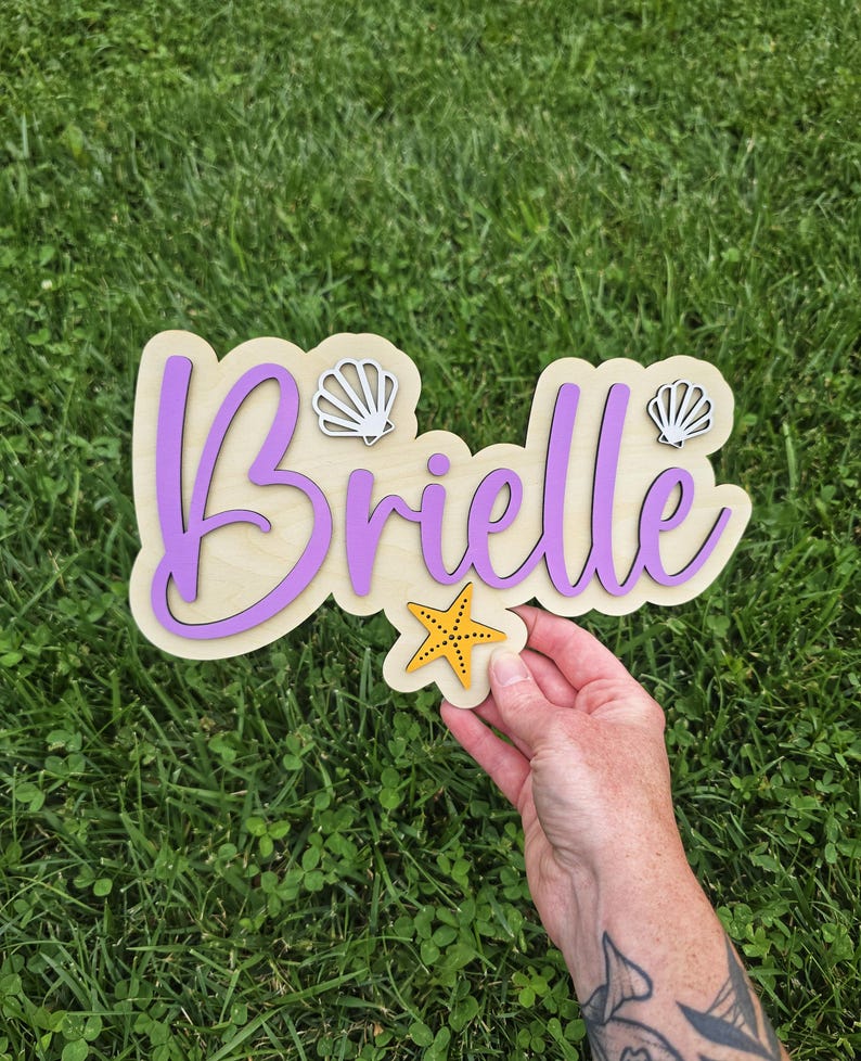 May include: Wooden sign with the name "Brielle" in purple script, outlined in a darker purple, on a light wood base. The sign is decorated with two white seashell cutouts and a yellow starfish. The sign is held over green grass.