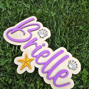 May include: Wooden sign with the name "Brielle" in purple script, accented with a yellow starfish and two seashell cutouts. The sign is on a light-colored wooden base with a scalloped edge.