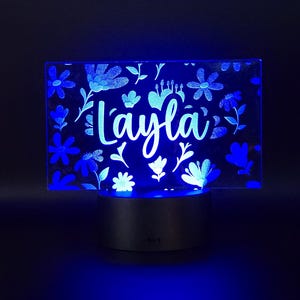 May include: A blue LED night light with the name "Layla" in a cursive font. The light is rectangular with a floral design and sits on a silver base. The flowers and leaves are in a darker blue.