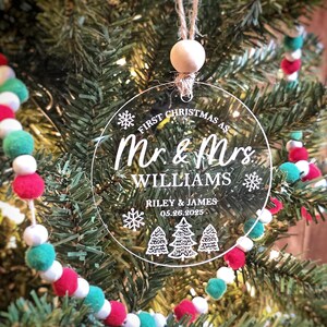 May include: Clear, round Christmas ornament with the text "First Christmas as Mr. & Mrs. Williams" and the names Riley & James, with the date 05.26.2025. The ornament hangs from a twine loop with a wooden bead, set against a Christmas tree.