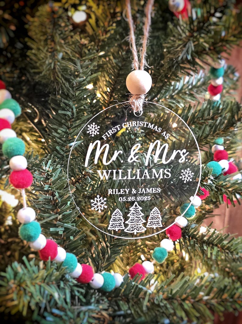 May include: Clear, round Christmas ornament with the text "First Christmas as Mr. & Mrs. Williams" and the names Riley & James, with the date 05.26.2025. The ornament hangs from a twine loop with a wooden bead, set against a Christmas tree.