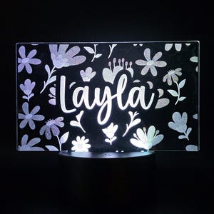 May include: A rectangular LED night light with the name "Layla" in a cursive font. The light is surrounded by a floral pattern with various flower and leaf designs. The light is on a black base.