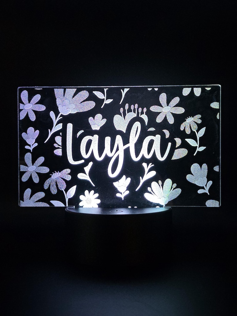 May include: A rectangular LED night light with the name "Layla" in a cursive font. The light is surrounded by a floral pattern with various flower and leaf designs. The light is on a black base.