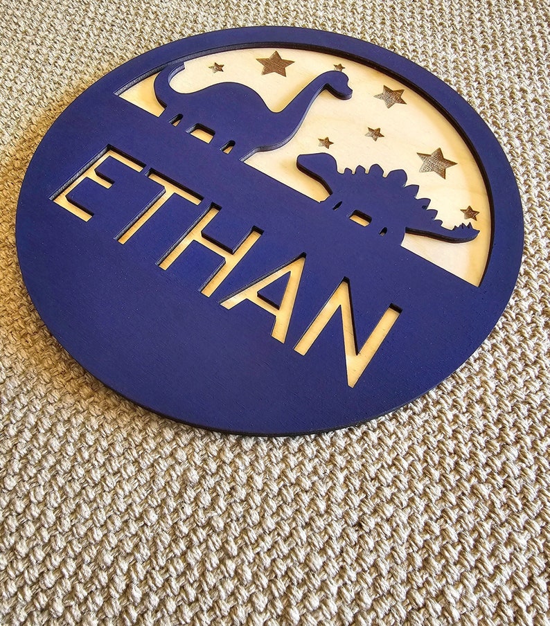 May include: A blue wooden circle with the name "ETHAN" cut out in the center. The circle has a wood veneer cutout of two dinosaurs and gold stars on a white background.