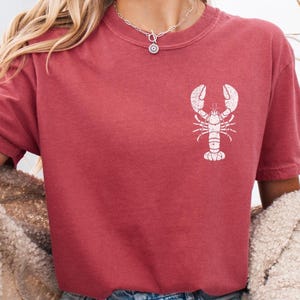 以下が含まれることがあります： 白いロブスターのグラフィックがプリントされた赤いTシャツを着た女性。Tシャツはクルーネックで半袖。女性はデニムのショートパンツと白いカーディガンを着ています。