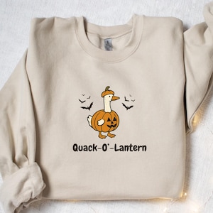 May include: Sand-colored sweatshirt with a Halloween-themed graphic. The design features a cartoon duck dressed as a jack-o'-lantern, with the text "Quack-O'-Lantern" below. Black bat cutouts are scattered around the sweatshirt.