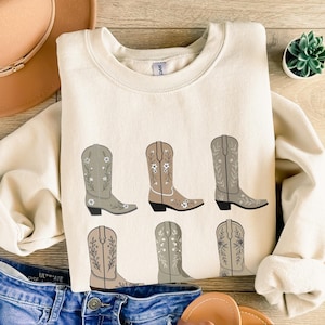 May include: A cream colored sweatshirt with a graphic of six brown cowboy boots with floral embroidery. The boots are arranged in two rows of three.