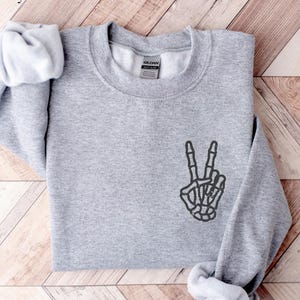 Könnte beinhalten: Sportgraues Sweatshirt mit Rundhalsausschnitt und langen Ärmeln. Das Sweatshirt hat eine schwarze Skeletthand, die ein Friedenszeichen auf der Vorderseite zeigt. Der Text "Sport Grey" befindet sich in der oberen linken Ecke.