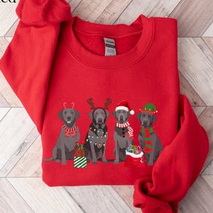 May include: Red sweatshirt featuring a festive design of four gray Labrador Retrievers. Each dog is adorned with holiday accessories like antlers, Santa hats, and scarves. The word "Red" is in the top left corner.