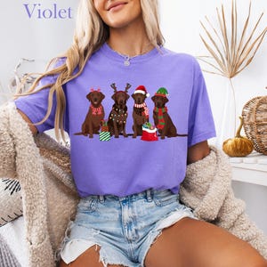 Op de afbeelding: Een lila T-shirt met een afbeelding van vier bruine Labradors in kerstoutfits. De honden dragen feestelijke accessoires zoals kerstmutsen en sjaals. Het woord "Violet" staat bovenaan het shirt.