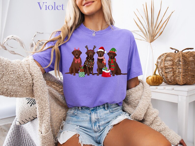 Op de afbeelding: Een lila T-shirt met een afbeelding van vier bruine Labradors in kerstoutfits. De honden dragen feestelijke accessoires zoals kerstmutsen en sjaals. Het woord "Violet" staat bovenaan het shirt.