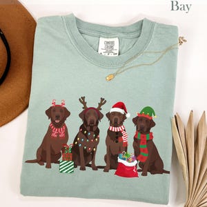 Op de afbeelding: Een lichtgroen Comfort Colors T-shirt met een afbeelding van vier bruine Labrador Retrievers met kerstaccessoires. Elke hond heeft een andere feestelijke versiering, waaronder een kerstmuts, een elfenmuts en rendiergewei. Het woord "Bay" staat bovenaan.