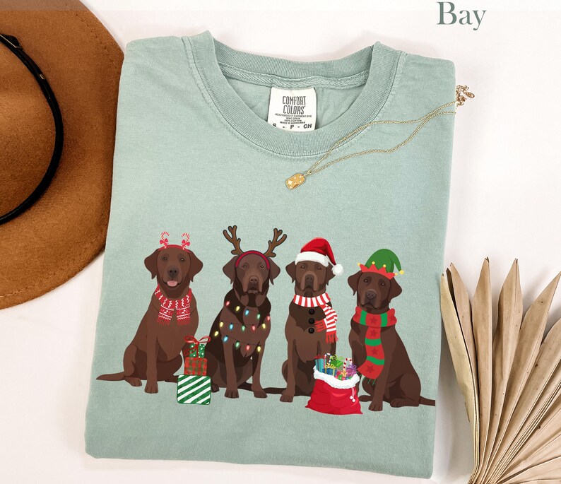 Op de afbeelding: Een lichtgroen Comfort Colors T-shirt met een afbeelding van vier bruine Labrador Retrievers met kerstaccessoires. Elke hond heeft een andere feestelijke versiering, waaronder een kerstmuts, een elfenmuts en rendiergewei. Het woord "Bay" staat bovenaan.