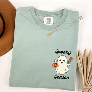 May include: A light blue t-shirt with a white ghost holding a pumpkin and a flower. The text "Spooky Season" is printed in black below the ghost.