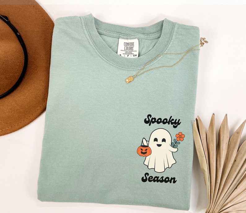 May include: A light blue t-shirt with a white ghost holding a pumpkin and a flower. The text "Spooky Season" is printed in black below the ghost.